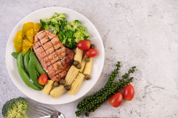 Chicken steak topped with white sesame, peas, tomatoes, broccoli and pumpkin in a white plate.