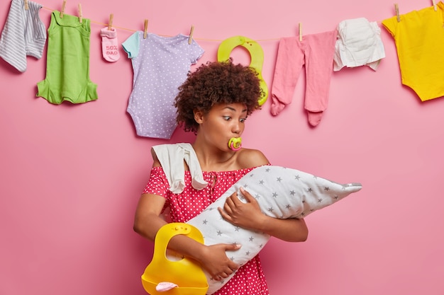 Child care, motherhood concept. Busy curly haired mother embraces newborn, poses with baby accessories, busy nursing child