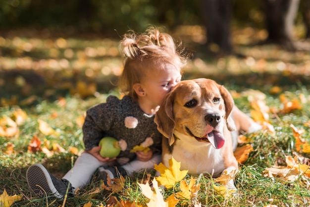 Free photo child girl kissing her dog sitting in grass at forest