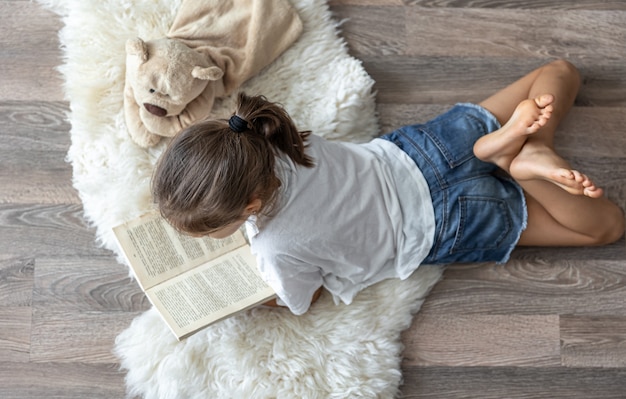 Free photo the child reads a book lying on a cozy rug at home with his favorite toy teddy bear.