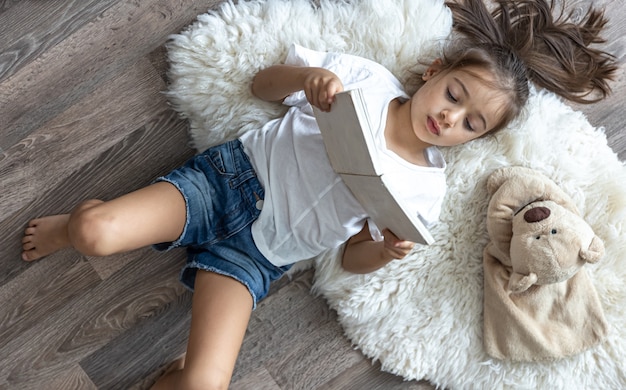 The child reads a book lying on a cozy rug at home with his favorite toy teddy bear.