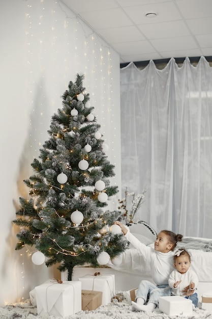 Free photo child in a white sweater. daughters stitting near christmas tree.two sisters at home.