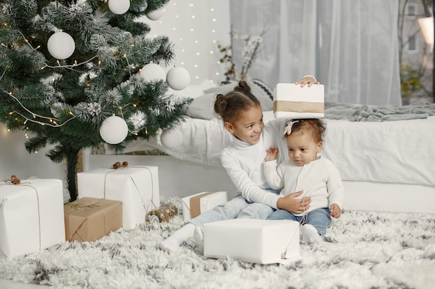 Child in a white sweater. Daughters stitting near Christmas tree.Two sisters at home.