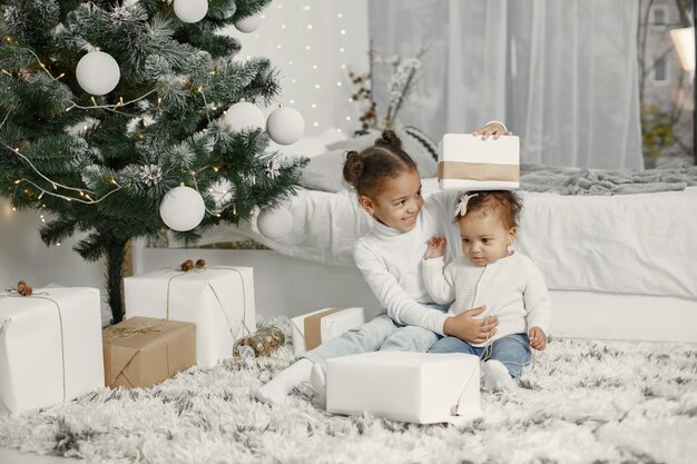 Child in a white sweater. Daughters stitting near Christmas tree.Two sisters at home.