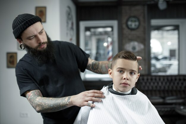 Children hairdresser cutting little boy against a dark background. Contented cute preschooler boy getting the haircut. The hand of the master have tattoo with the word shave