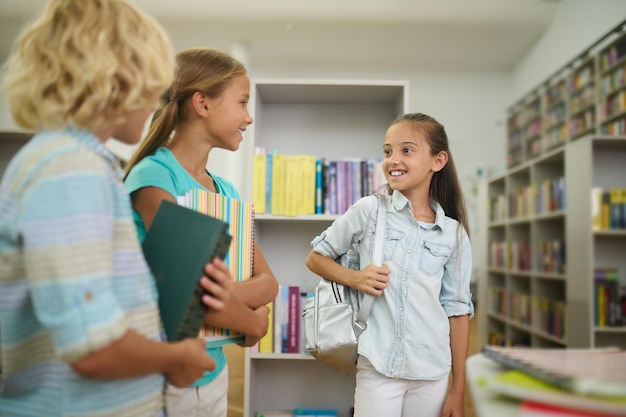 Children, library. Two smiling long-haired girls with backpack and notebooks and blonde boy with books are standing talking in light room in library