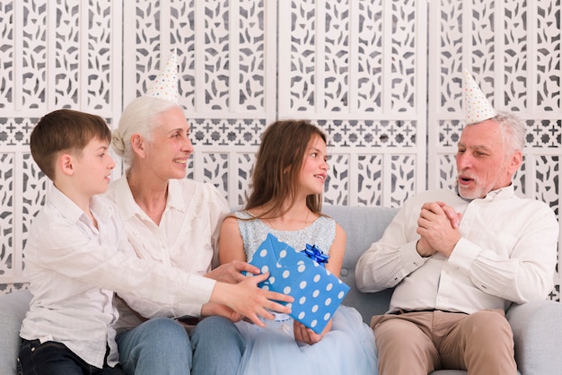 Children with grandmother giving gift to their happy grandfather sitting on sofa