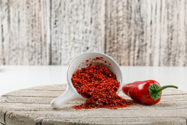 Chili powder with red pepper and wooden piece in a white scoop on white and wooden grunge wall, side view.