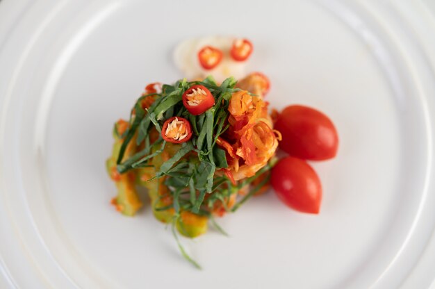 Chilies, kaffir lime leaves, bitter gourd and tomatoes stacked together on a white plate.