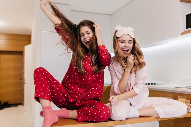 Chilling girl in pink socks sitting on wooden table. Indoor photo of cute young woman in pink sleepwear having fun in kitchen.