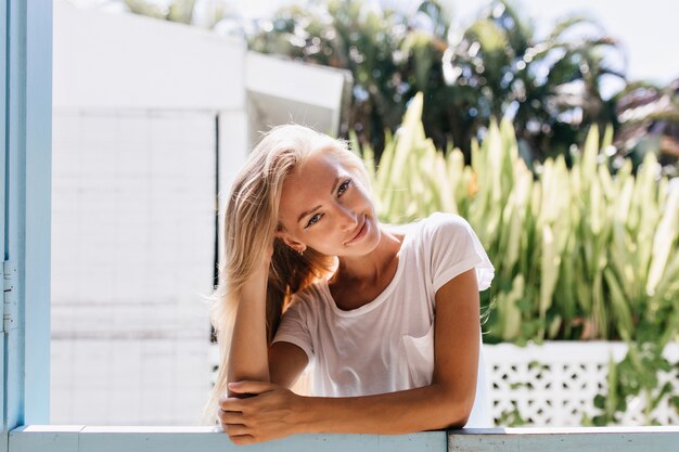 Chilling tanned woman posing with sly smile. Fascinating young lady with blonde shiny hair enjoying weekend.