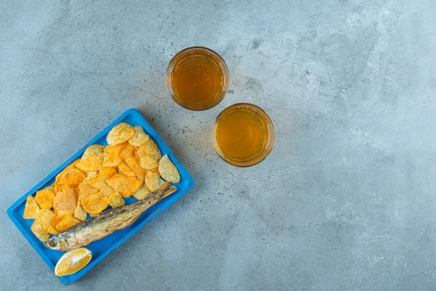 Chips and fish on a wooden plate next to two glasses of beers , on the marble table. 