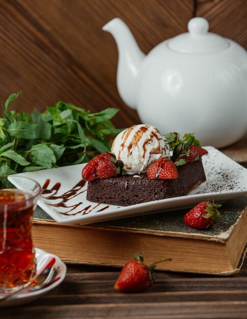 Chocolate brownie with icecream ball and strawberries , and a glass of tea 