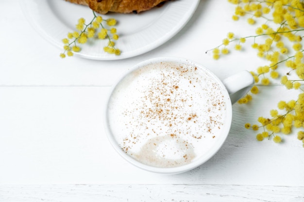 Chocolate croissant and cappuccino decorated with mimosa flowers on a white table Spring Mother's Day concept