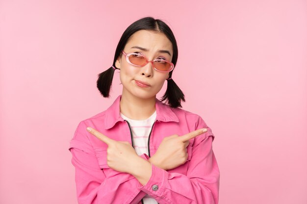 Choice Stylish korean girl asian female model points fingers sideways shows two variants product advertisement demonstrating items standing over pink background