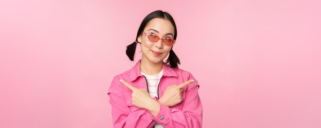 Choice Stylish korean girl asian female model points fingers sideways shows two variants product advertisement demonstrating items standing over pink background