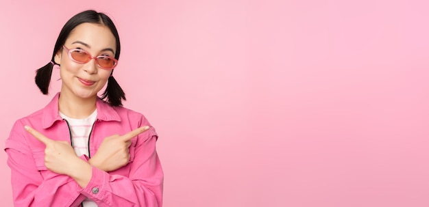 Choice Stylish korean girl asian female model points fingers sideways shows two variants product advertisement demonstrating items standing over pink background