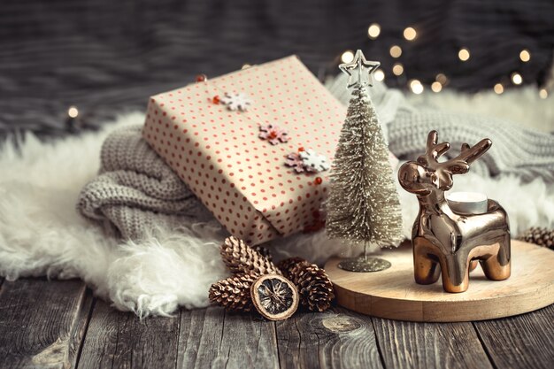 Christmas festive background with toy deer with a gift box, blurred background with golden lights, festive background on wooden deck table and cozy sweater on background