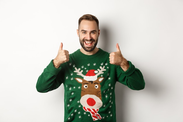 Free photo christmas, holidays and celebration. happy young man enjoying new year party, showing thumbs up in approval, like something good, standing over white background