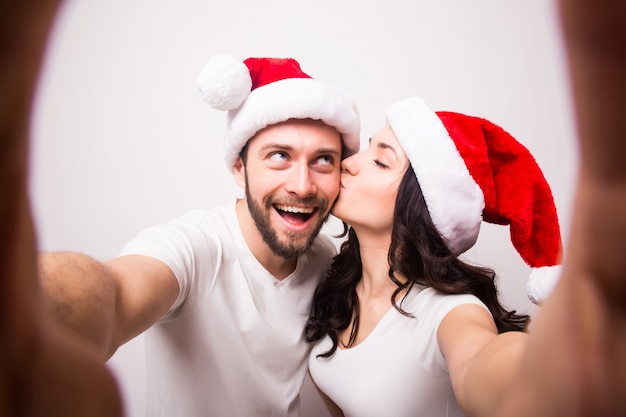 Free photo christmas, holidays, technology and people concept - happy couple in santa hats taking selfie picture from hands on white background. they look at camera and kiss