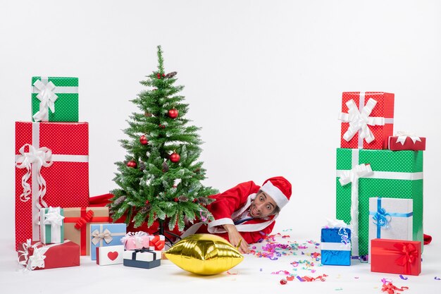 Christmas mood with santa claus lying behind christmas tree near gifts in different colors on white background