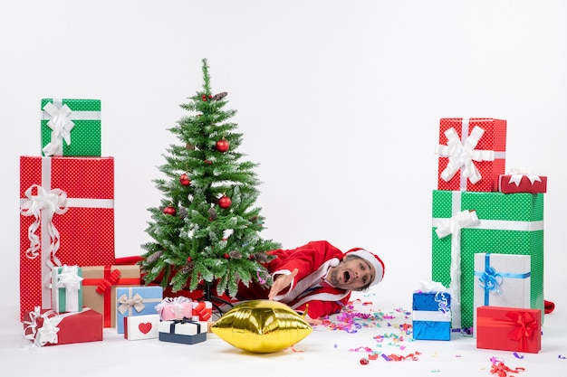 Christmas mood with young sad santa claus lying behind christmas tree near gifts in different colors on white background