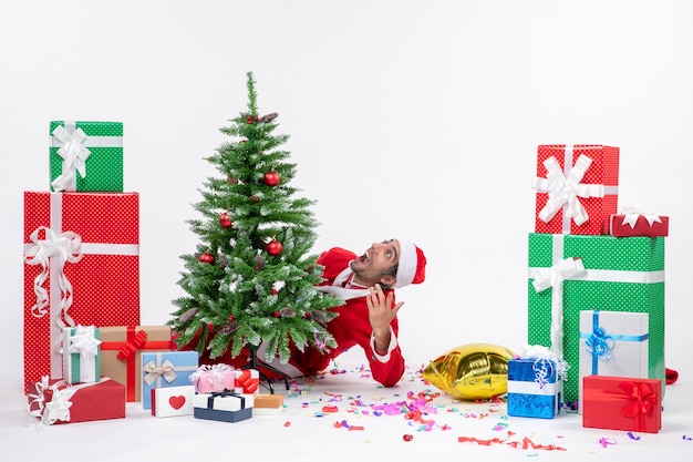 Christmas mood with young santa claus hiding behind christmas tree near gifts in different colors on white background