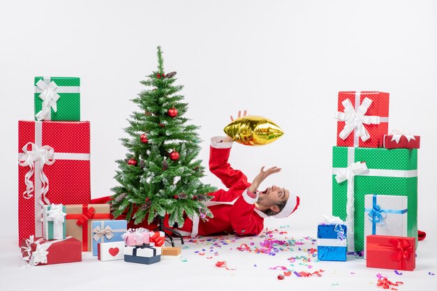 Christmas mood with young santa claus lying behind christmas tree near gifts in different colors on white background