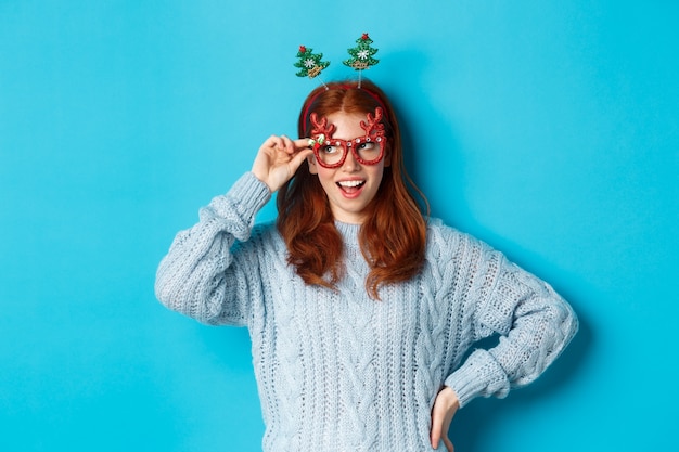 Christmas party and celebration concept. Cute redhead teen girl celebrating New Year, wearing xmas tree headband and funny glasses, looking left amused, blue background.