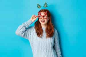 Free photo christmas party and celebration concept. cute redhead teen girl celebrating new year, wearing xmas tree headband and funny glasses, looking left amused, blue background