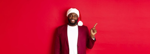 Free photo christmas party and holidays concept handsome black man in santa hat pointing finger left showing ad