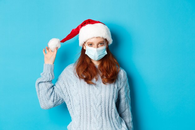 Christmas, quarantine and covid-19 concept. Redhead girl wearing face mask and playing with santa hat, celebrating New Year on lockdown, standing over blue background.