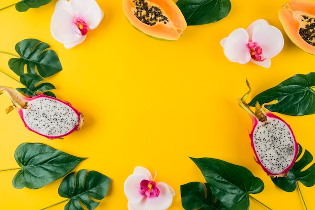 Free photo circular frame made with artificial leaves; orchid flower; papaya and dragon fruits on yellow backdrop