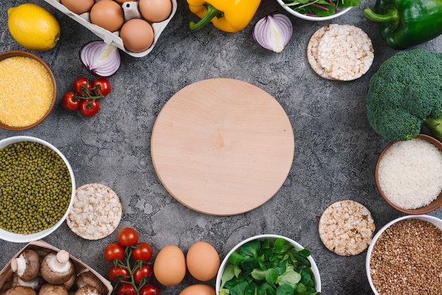 Circular wooden chopping board surrounded with fresh vegetables and eggs on concrete background