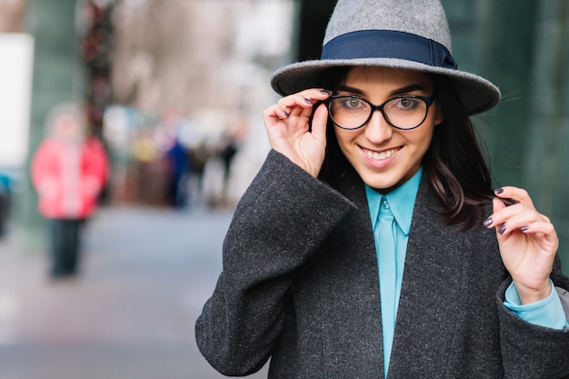 City portrait stylish charming young woman in grey coat, hat walking on street. Modern black glasses, smiling, expressing true happy emotions, luxury lifestyle, fashionable model.