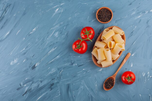 A clay board of dry raw tube pasta with fresh red tomatoes and pepper corns .