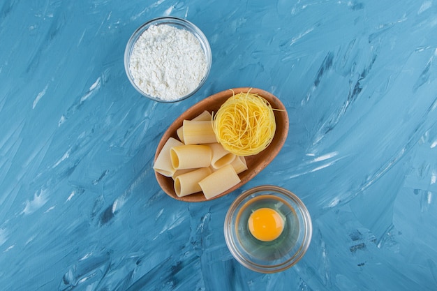 A clay board of nest noodles and a glass bowl of flour on a blue background. 