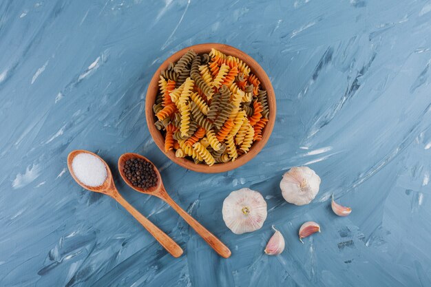 A clay bowl of multi colored raw pasta with garlics on a blue table.