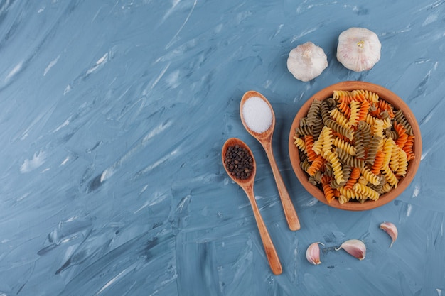 A clay bowl of multi colored raw pasta with garlics on a blue table.