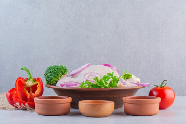 Free photo clay bowl of raw whole chicken with various condiments on stone surface.