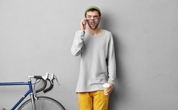 Clever sportsman looking through big glasses with serious expression, drinking delicious hot coffee, going to competitions or in gym.