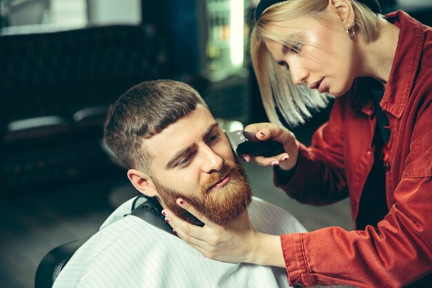 Client during beard shaving in barbershop. Female barber at salon. Gender equality. Woman in the male profession.