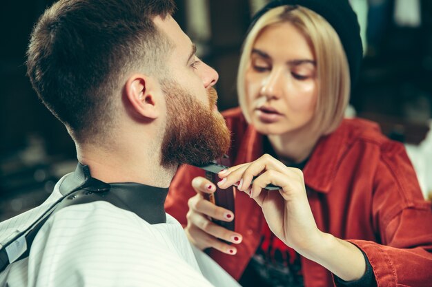 Client during beard shaving in barbershop. Female barber at salon. Gender equality