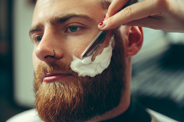 Free photo client during beard shaving in barbershop. female barber at salon. gender equality. woman in the male profession. hands close up