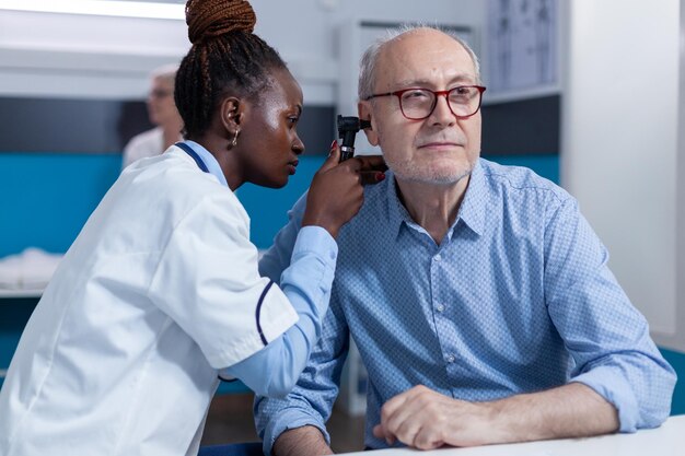 Clinic otology specialist consulting senior patient using otoscope to check ear infection. Hospital otologist examining sick retired man internal ear condition while in doctor cabinet.