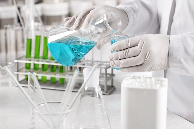 Clinical worker dressed in white gown and gloves holding glass beakers with blue liquid