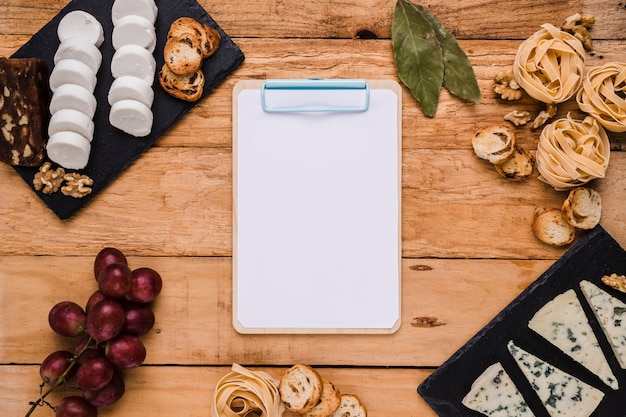 Clipboard with blank paper surrounded with tasty cheese and ingredients over textured wood