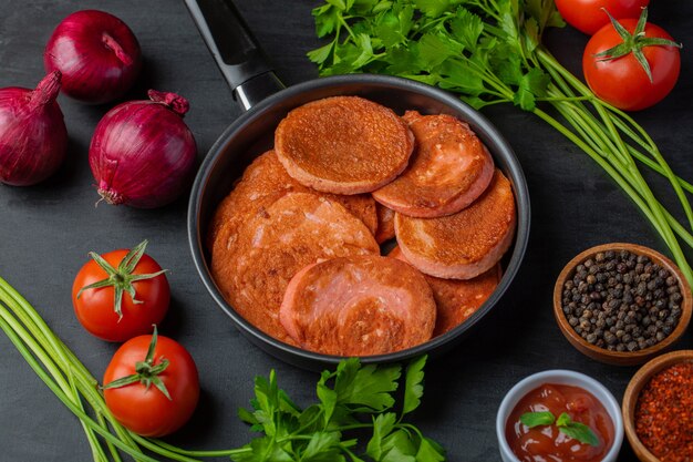 Clos up photo of fried salami slices in the frying pan and various kind of vegetables around. 