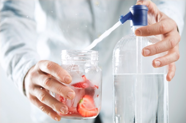 Close focus bartender makes homemade lemonade, pours sparkling water to rustic jar with strawberry slices and ice cubes inside