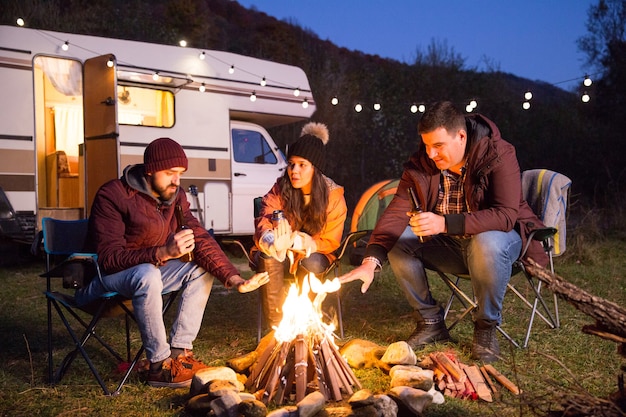 Free photo close friends drinking beer together in the mountains and warming their hands around camp fire. retro camper van with light bulbs.